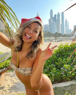 Woman in a Santa hat and bikini making a peace sign in front of skyscrapers.