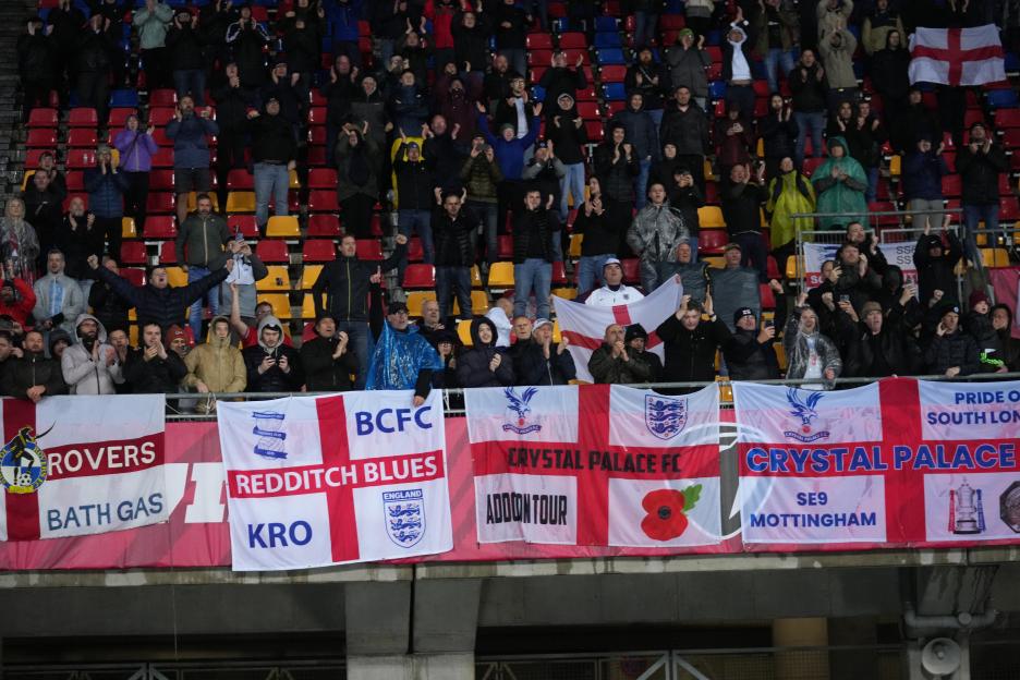 Football fans at a stadium cheering, with banners for various football clubs and England flags hanging below them.