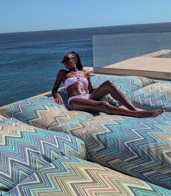 AJ Odudu relaxing on patterned cushions with the ocean in the background.