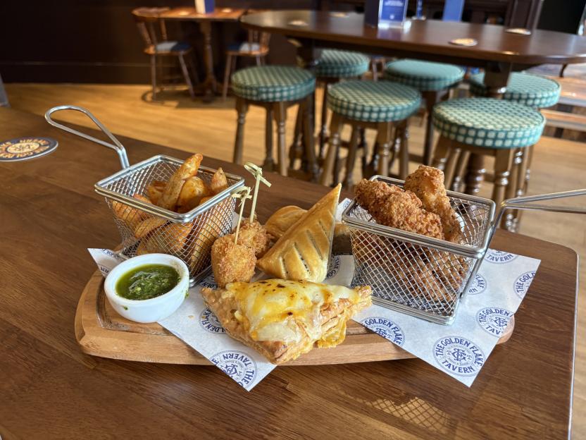 Assorted pub snacks, including crispy chicken, wedges, pastries, and a small dish of green sauce, served on a wooden board at The Golden Flake Tavern.
