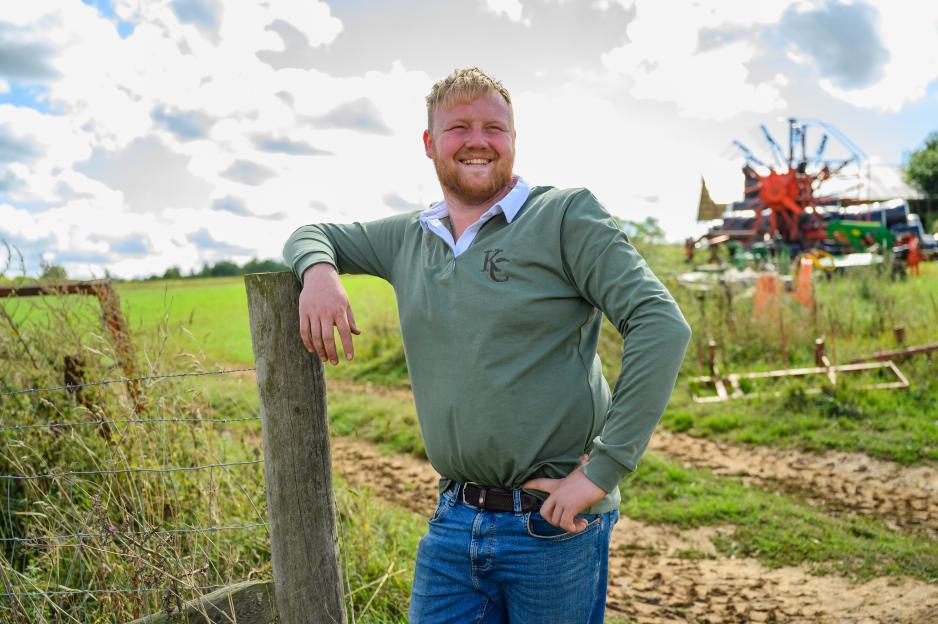 Kaleb Cooper, a man with a reddish beard, smiling and leaning on a metal gate in a barn, with cows behind him.