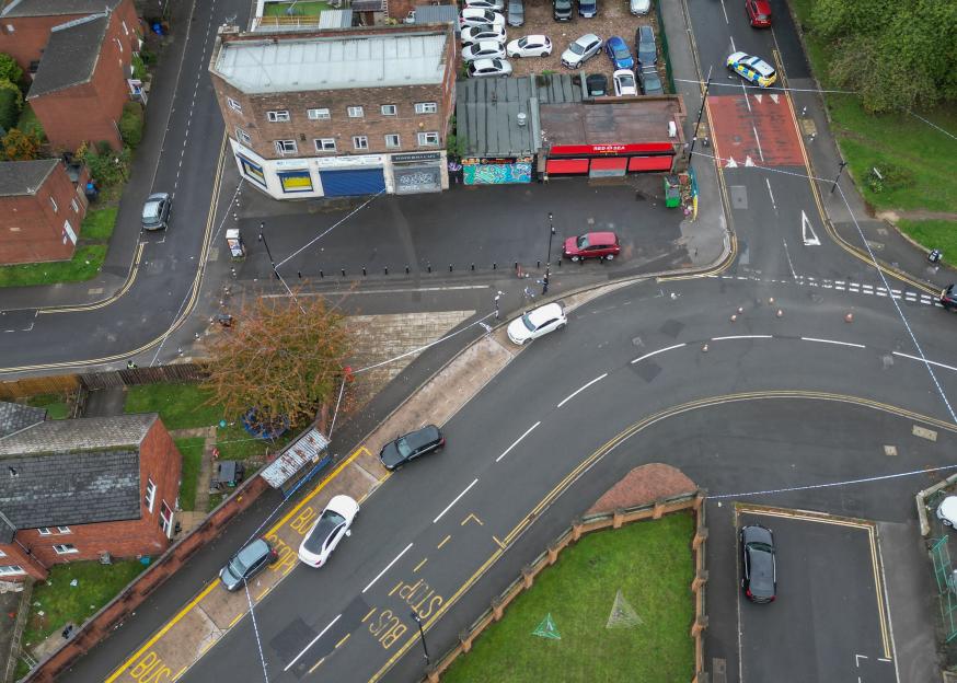Aerial view of a crime scene in Sheffield, UK, with police tape cordoning off a street with several parked cars and buildings, including a "Red Sea" shop.