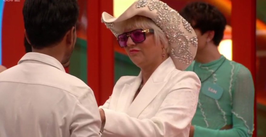 A person in a white suit, embellished cowboy hat, and purple sunglasses talks to another person, with a third person wearing a green top and a name tag that reads "SAM" in the background.