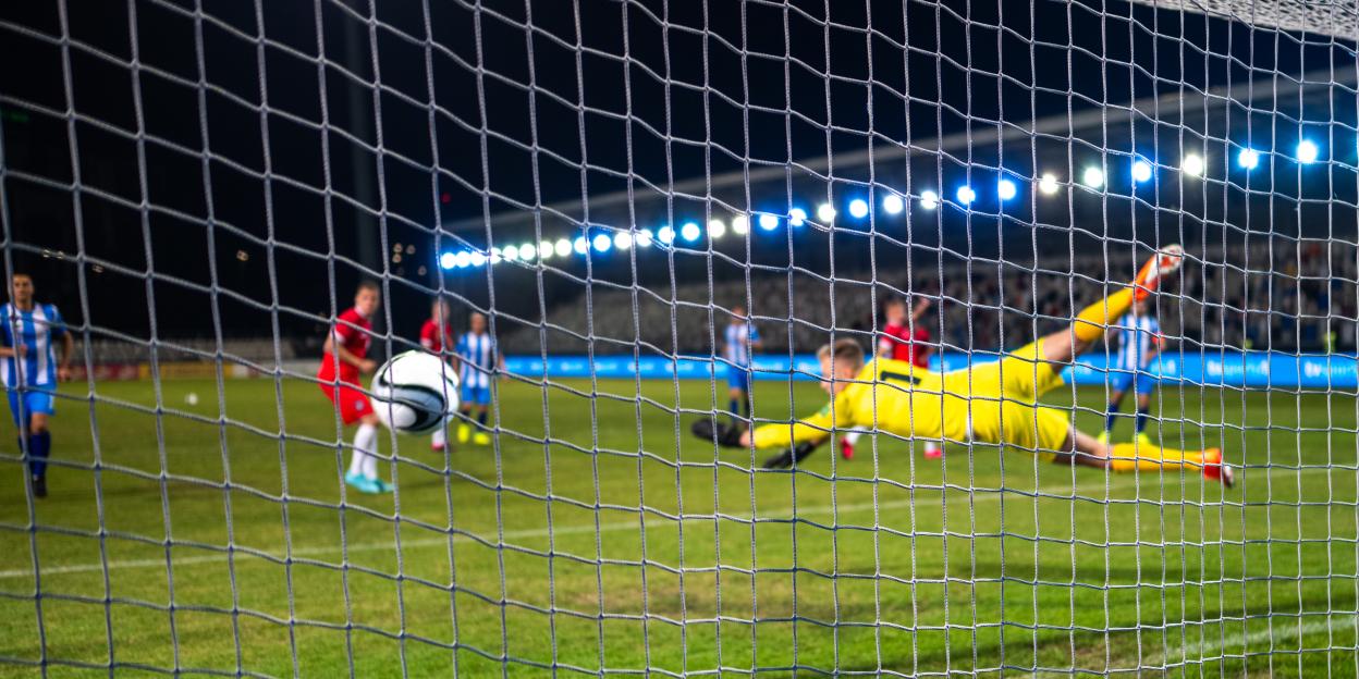 A soccer goalkeeper in yellow diving to catch the ball as seen through the goal net.