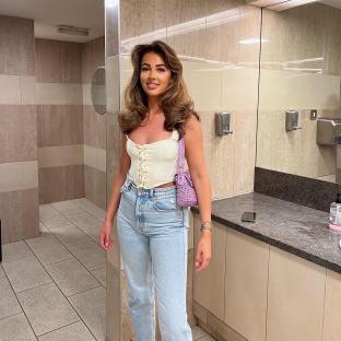 Woman in a cream corset top and light blue jeans in a restroom.