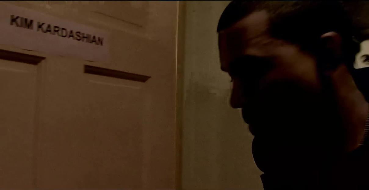Kanye West looking toward a door with a "Kim Kardashian" sign, with a man in the background.