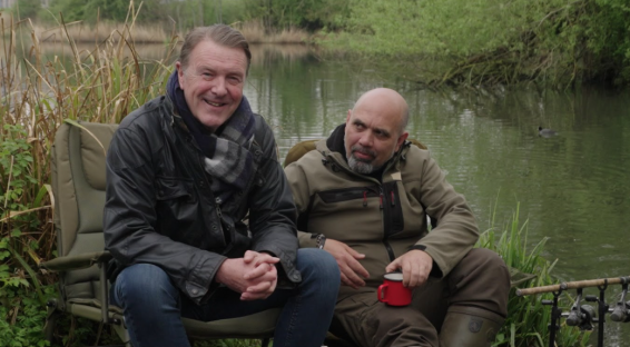 Two men sitting in chairs by a lake, holding mugs.