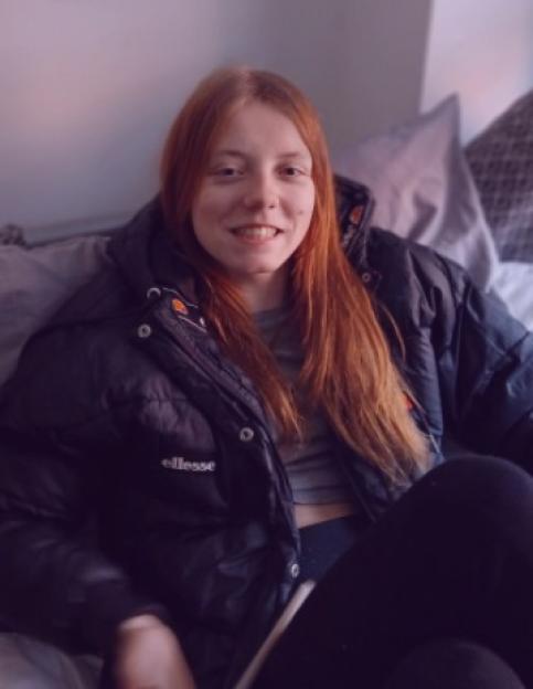 Woman with long red hair smiling while sitting on a bed in a black jacket.