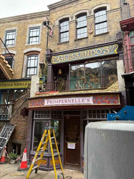 A behind-the-scenes view of the Diagon Alley set for the Harry Potter television series, showcasing buildings for Glissando's, Primpernella's, and Antonia Abraxas's, with scaffolding and a ladder in the foreground.