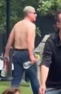 Man holding papers and talking while a shirtless man walks in the background.