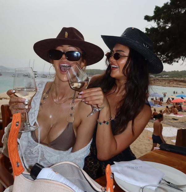 Two women in hats and sunglasses drinking wine at a beachside restaurant.