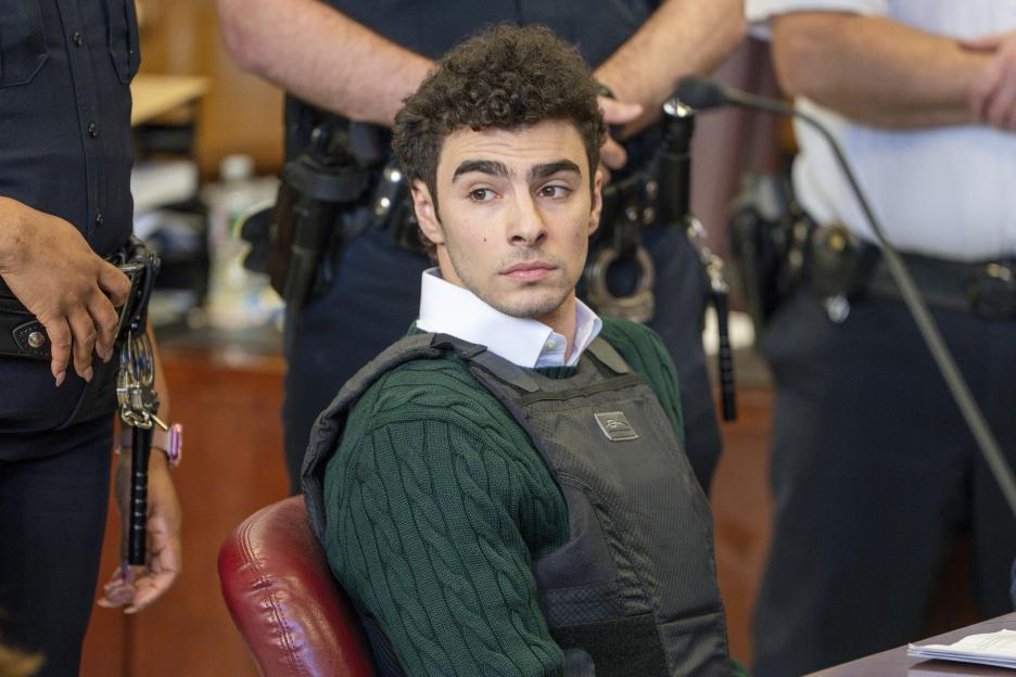 Luigi Mangione, accused of fatally shooting the UnitedHealthcare CEO, sits in court wearing a green sweater and a dark-colored ballistic vest, with police officers standing behind him.