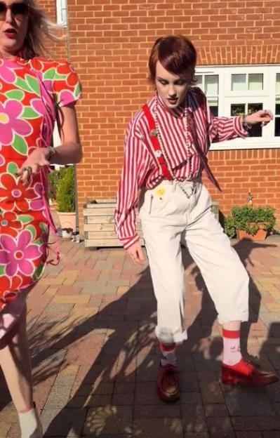 Two women dancing outdoors; one in a red and pink floral dress, the other in a red and white striped shirt and cream pants.