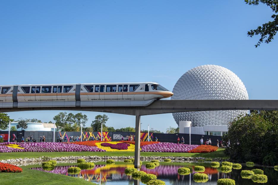 A monorail zips past flower displays at Epcot with Spaceship Earth in the background.