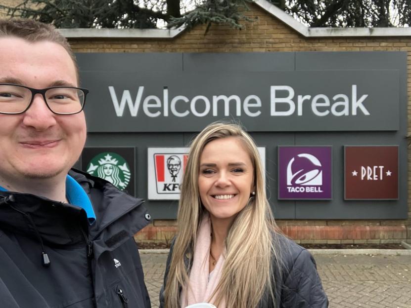Rich James Cross and another person smiling in front of a "Welcome Break" sign featuring logos for Starbucks, KFC, Taco Bell, and Pret.