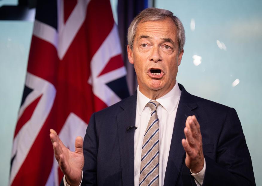 Nigel Farage speaking at a podium with two Union Jack flags behind him.