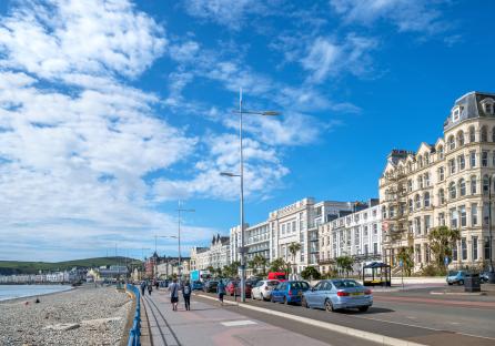 Pedestrians walking along a promenade with parked cars and hotels in Douglas, Isle of Man.