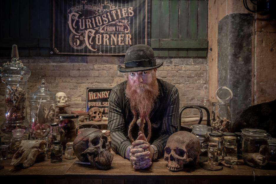 A man with a long red beard sits behind a table displaying skulls and jars filled with bones and other curiosities at a satanic flea market.