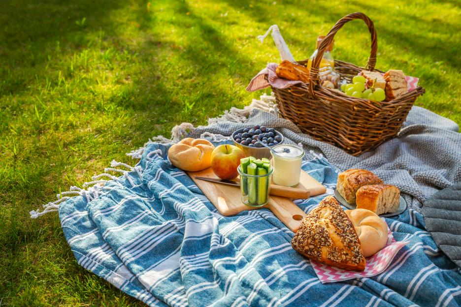 Picnic blanket with basket, bread, fruit, and yogurt on the grass.