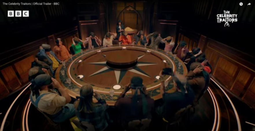 A circular table of contestants in a room with a moderator standing in the back.