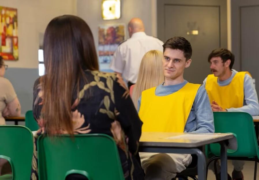 A man in a yellow vest sits at a table talking to a woman. Other people are in the background.