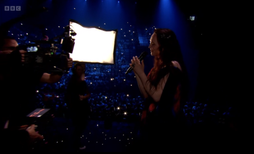 Singer performing on stage with a large audience and camera crew.