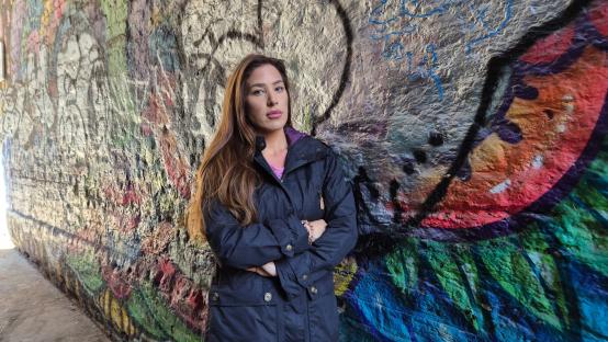 Woman with arms crossed standing in front of a graffiti-covered wall.