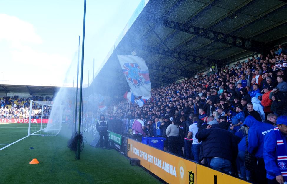A football stadium crowd being sprayed by water from pitchside sprinklers, with a large flag visible in the foreground.