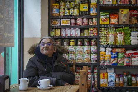 A coffee shop customer sits at a table with a cup of coffee, in front of shelves stocked with various food items.