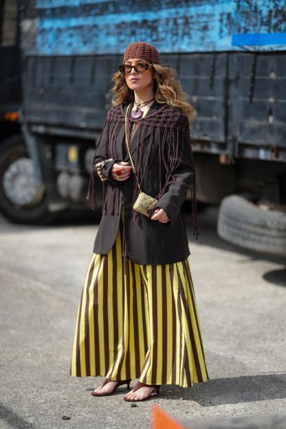 Woman wearing a brown macramé capelet, striped trousers, and layered jewelry.
