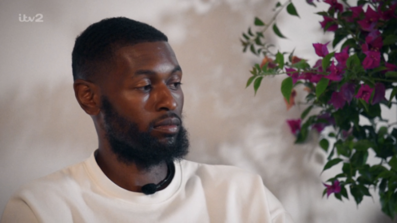 A man with a beard, wearing a white crewneck shirt with a lavalier microphone attached, sitting and looking thoughtfully to the right.