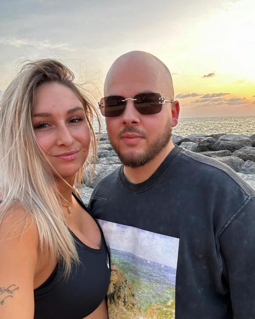 A man and woman posing for a selfie on a rocky shore with the ocean and sunset in the background.
