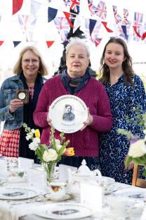 Three women at a table displaying a commemorative plate honoring Arnold Sharrocks, a WWII hero.