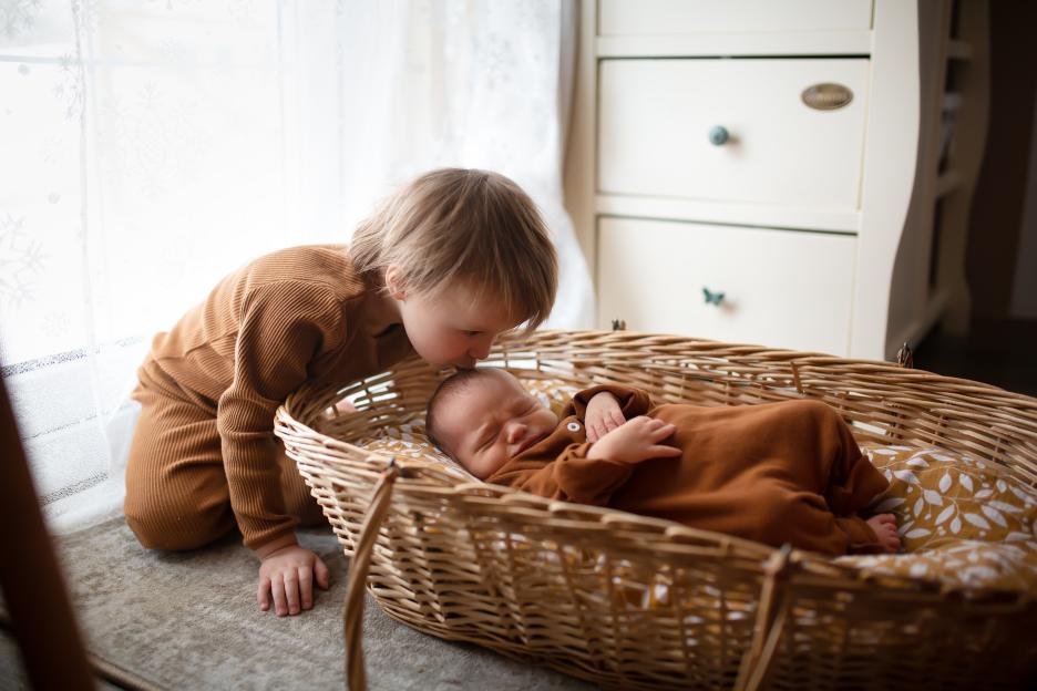 A toddler kissing a newborn baby in a wicker basket.