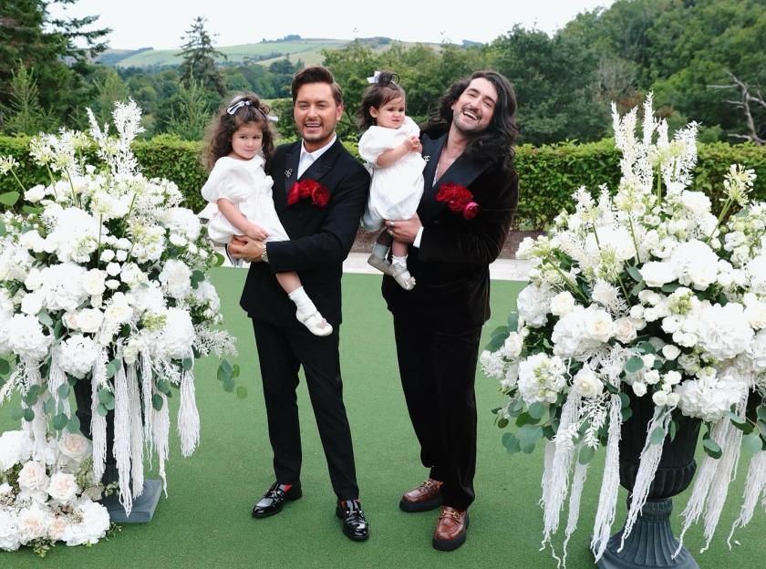 Two men in suits holding young children, standing between large white floral arrangements.