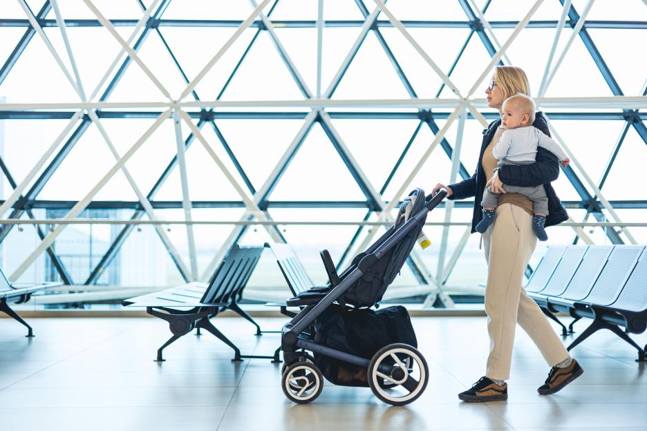 Mother carrying baby and pushing stroller through airport.