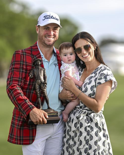 Golfer Justin Thomas, his wife Jillian Wisniewski, and their daughter Molly Grace smiling with the tournament trophy and plaid tartan jacket.