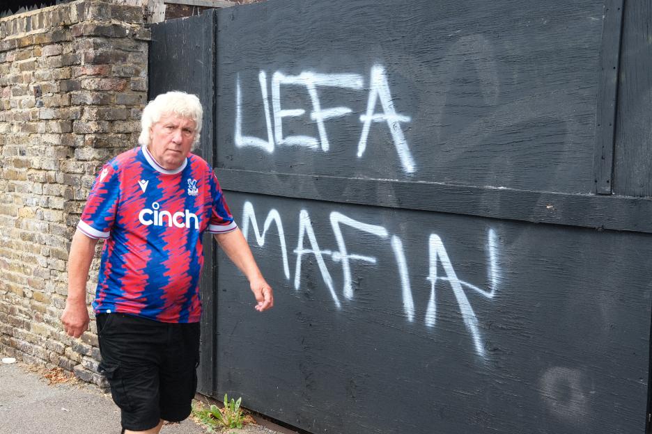 Graffiti reading "UEFA Mafia" on a wall, with a man in a Crystal Palace jersey walking by.