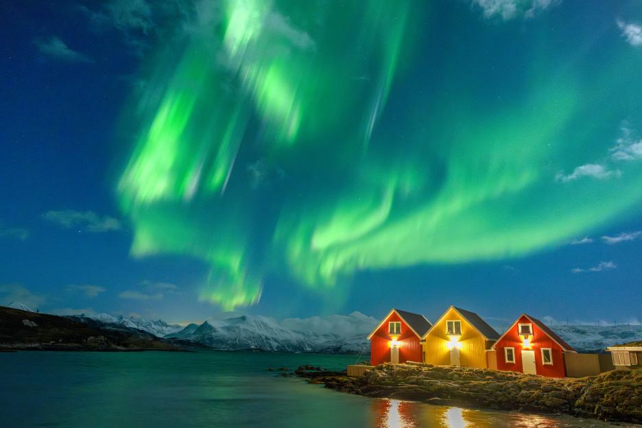 Aurora borealis over snowcapped mountains and cabins by the sea.