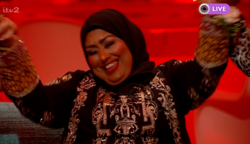 A person in a hijab and embroidered black top smiles broadly with arms raised.