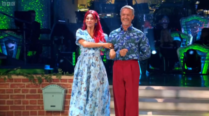 A woman with red hair in a blue floral dress and a man in a blue floral shirt and red pants standing arm in arm.
