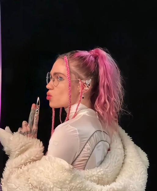 Grimes with pink braided hair and a high ponytail, wearing glasses and a white furry coat.