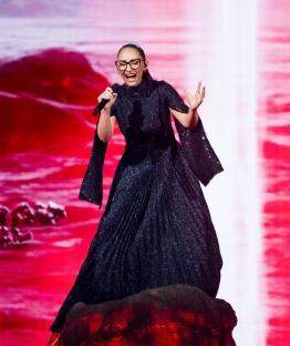 A woman in a sparkly black dress sings into a microphone on a stage with a red background.