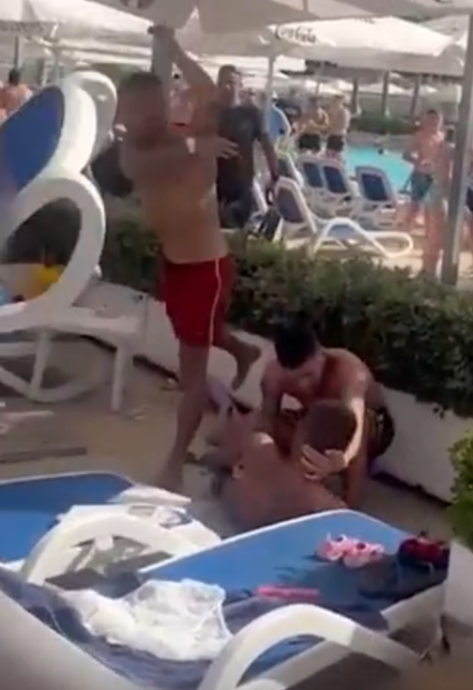 A man in red shorts punches another man who is kneeling on the ground next to an unconscious man, near a swimming pool.