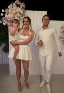 A woman in a white dress holds a toddler while standing next to a man in a white shirt and pants.