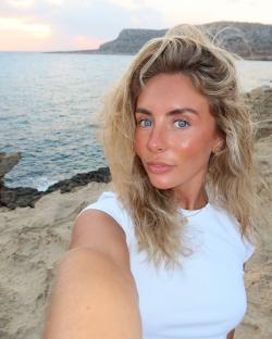 Selfie of a blonde woman wearing a white shirt near the ocean.