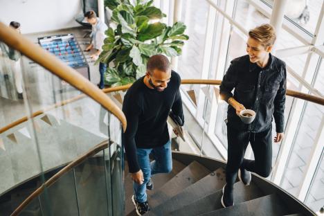Two smiling colleagues ascending an office staircase; one carries a laptop, the other holds a coffee cup.
