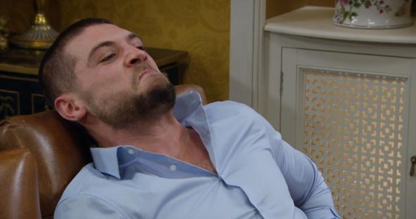 Caleb from Emmerdale in a light blue shirt, reclining in a chair.