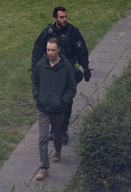 A man in a gray hoodie and tan shoes walks on a sidewalk with a police officer behind him.