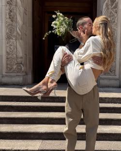 A man in beige trousers carrying his bride in a white tracksuit, kissing.